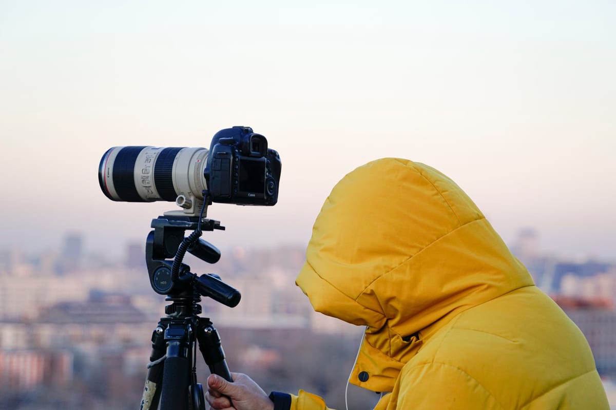 Fotografin in gelbem Parka hinter einer Spiegelreflexkamera mit Teleobjektiv auf einem Stativ über einer Stadtsilhouette