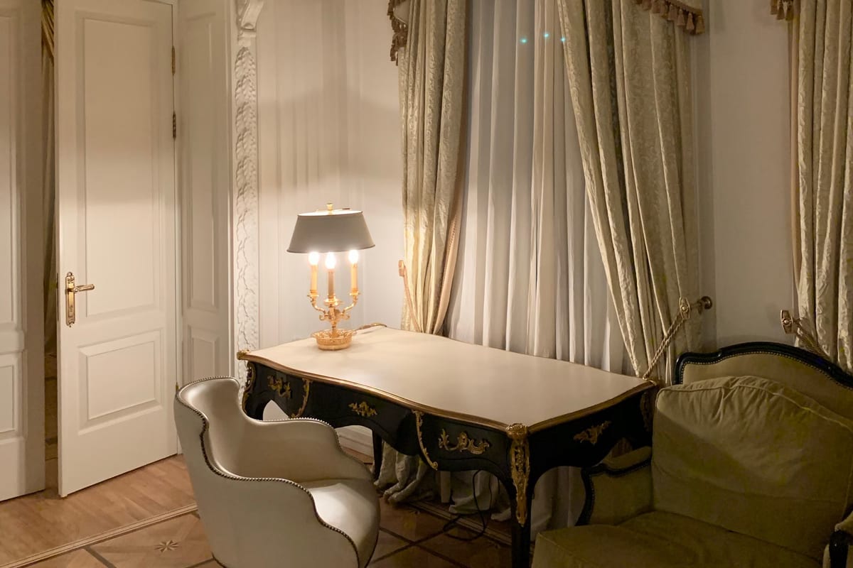 Salon einer historischen Hotelsuite mit Schreibtisch, Sessel, beleuchteter Tischlampe und schweren Vorhängen