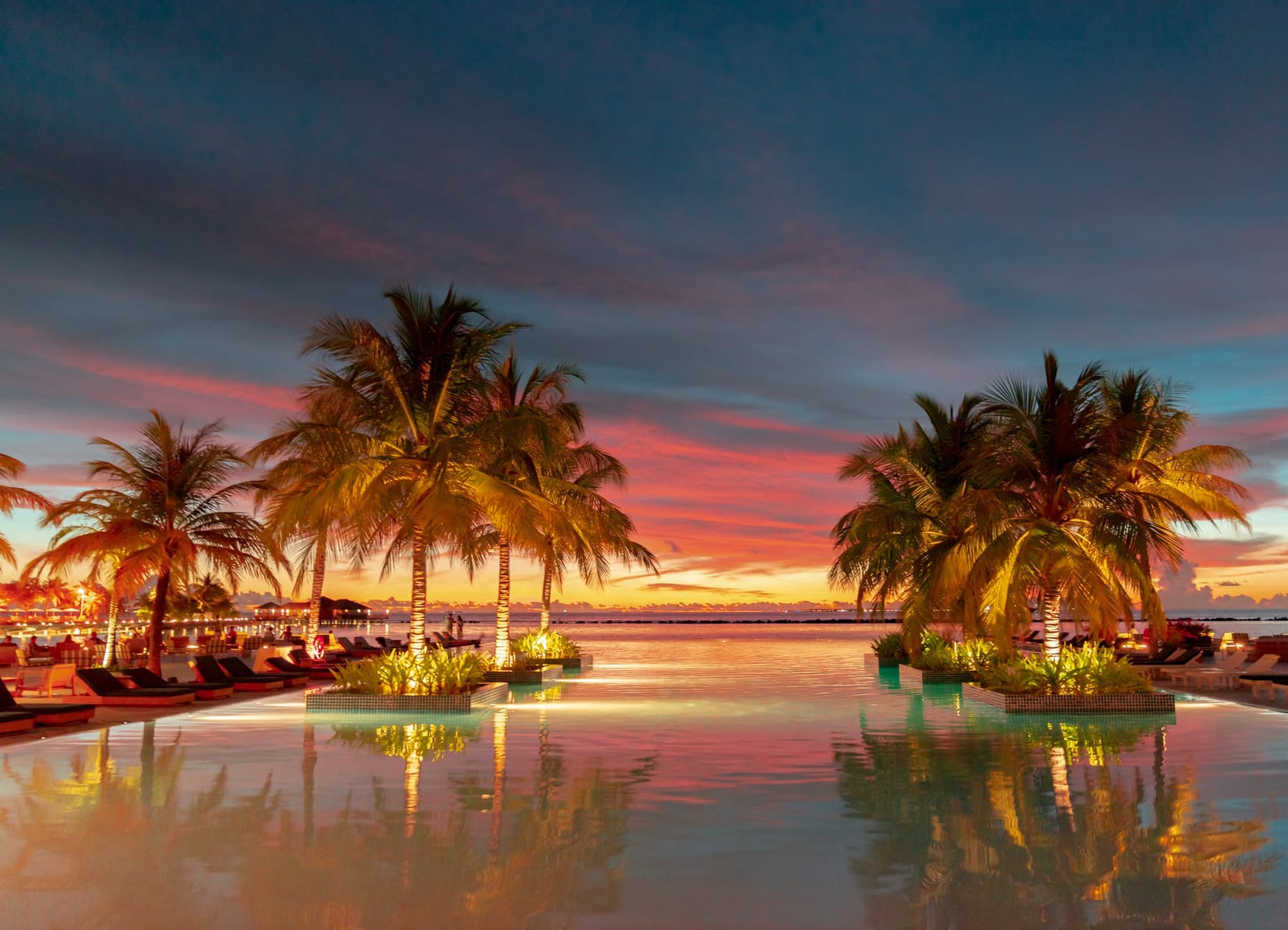 Infinity-Pool einer Strandanlage im Abendrot, beleuchtete Palmeninseln spiegeln sich im ruhigen Wasser